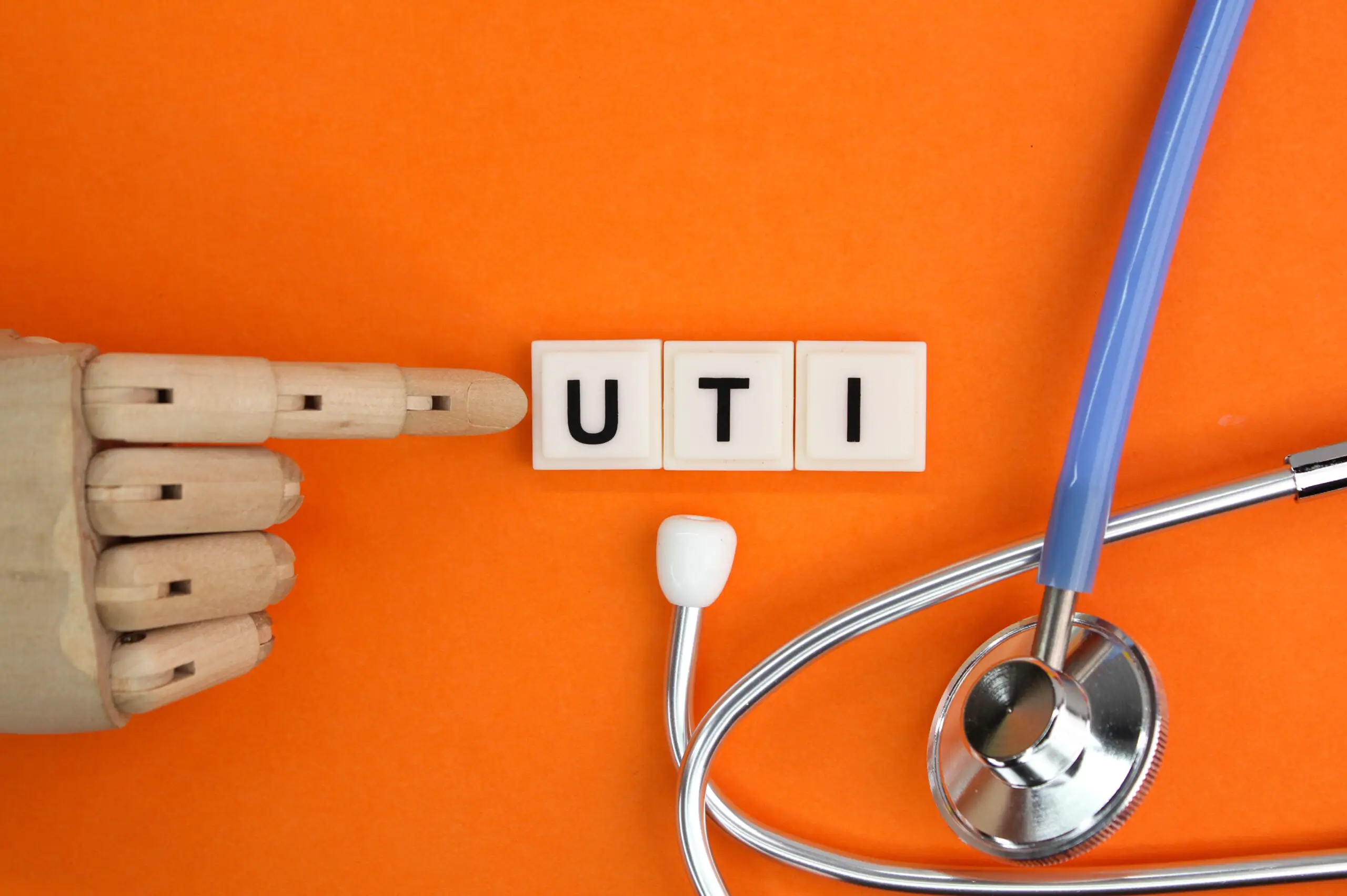 Wooden hand points to letter tiles spelling "UTI" near a stethoscope on orange.